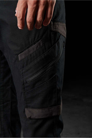 A close-up image shows a person's hand resting on their thigh, clad in FXD WP-11 pants. These black pants, crafted from 2-way stretch fabric, are highlighted by their multi-functional cargo pockets and multiple zippered compartments. The dark and blurred background accentuates the details of both the pants and the hand.