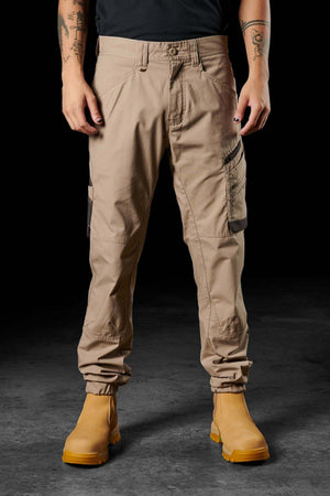 A person is standing against a dark background, wearing FXD WP-11 pants with multi-functional cargo pockets and light tan work boots. Only the lower half of their body, from waist to feet, is visible. The individual has tattoos on their arms and is wearing a dark shirt.
