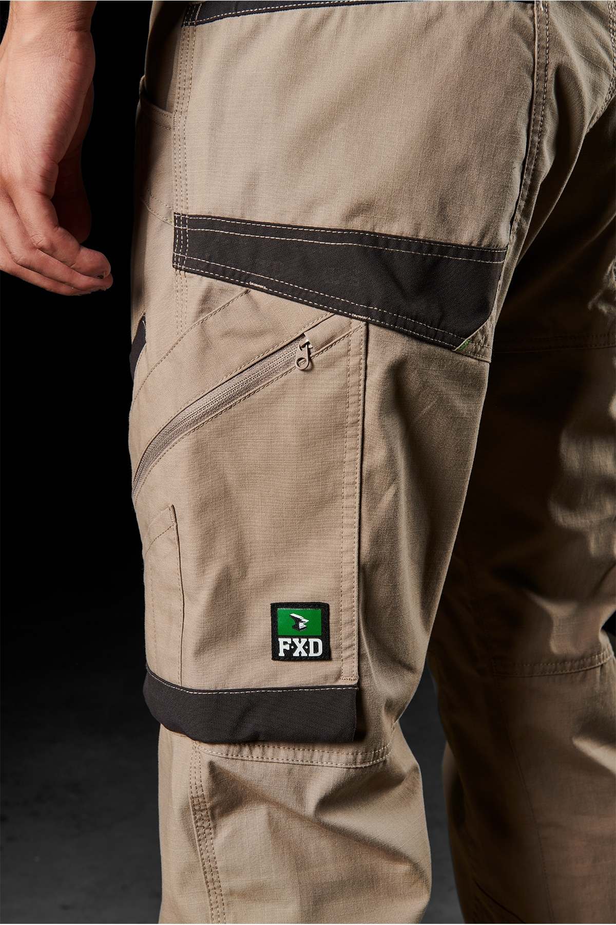 Close-up of a person wearing FXD WP-11 pants in beige with black accents and multi-functional cargo pockets. A green logo patch with "FXD" text is prominently displayed on the left cargo pocket. The 2-way stretch fabric provides comfort and flexibility, while the dark background highlights the details of the pants.