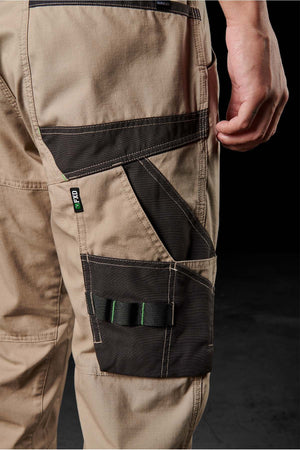 A close-up image of a person wearing beige FXD WP-11 work pants with black reinforced pockets and stitching. The pants, crafted from 2-way stretch fabric, feature various functional compartments, including a velcro pocket and multiple open tool slots on the side. The dark background accentuates the detailing of the pants.
