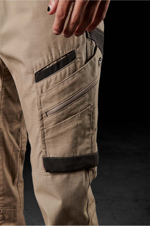 Close-up of a person's hand resting on their FXD WP-11 tan pants, highlighting the multi-functional cargo pockets, zippers, and black accents. The background is dark and out of focus.