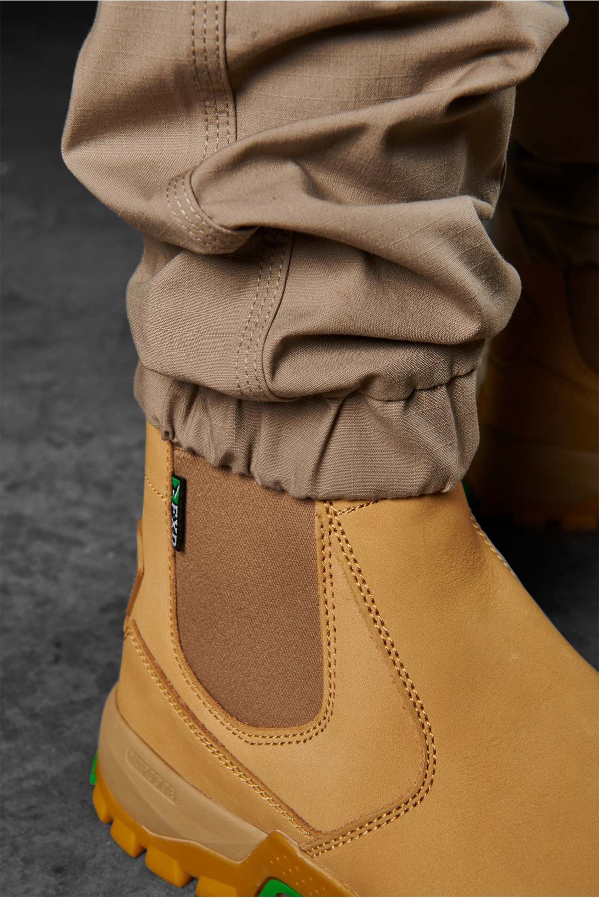 Close-up of a person wearing FXD WP-11 pants tucked into light brown work boots. The slightly wrinkled, tan FXD WP-11 pants feature multi-functional cargo pockets, and the boots have green accents on the soles. The background is a dark, textured surface.
