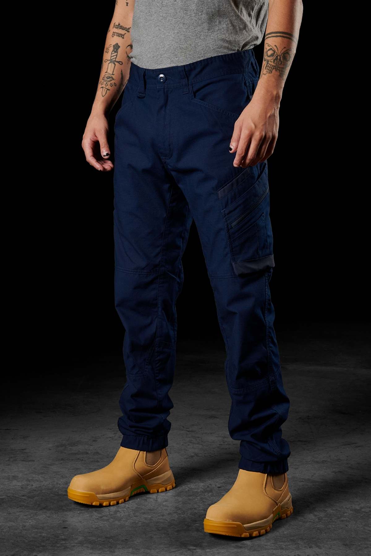 A person wearing FXD WP-11 pants in deep navy, showcasing the 2-way stretch fabric, stands against a dark backdrop. They are paired with a gray T-shirt and tan work boots. Tattooed arms hang relaxed by their sides, while the utility-rich cargo pockets complete the look.