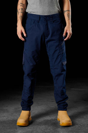 A person is shown from the waist down, wearing dark blue FXD WP-11 pants with cargo pockets. They are also sporting a grey shirt and tan work boots while standing against a black background. Tattoos are visible on both their forearms.
