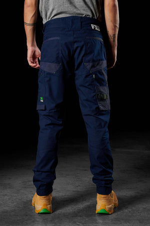 A person wearing dark blue FXD WP-11 work pants with multiple cargo pockets is viewed from the back. The pants feature a small logo on the rear waistband and have lighter blue accents. The individual is also wearing tan work boots and has tattoos on their arms.