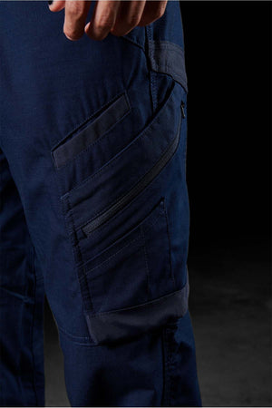 Close-up of a person's hand and a section of their FXD WP-11 pants in blue, highlighting multiple cargo pockets, including one with a zipper on the thigh. The dark background accentuates the details of the 2-way stretch fabric.