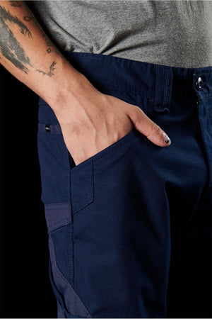 Close-up of a person's hand in the pocket of navy blue FXD WP-11 pants, showcasing the 2-way stretch fabric. The person is wearing a grey shirt and displaying a tattoo on their forearm. The cargo pocket features a flap and intricate stitching detail, and the person's short fingernails are painted. The background is black.