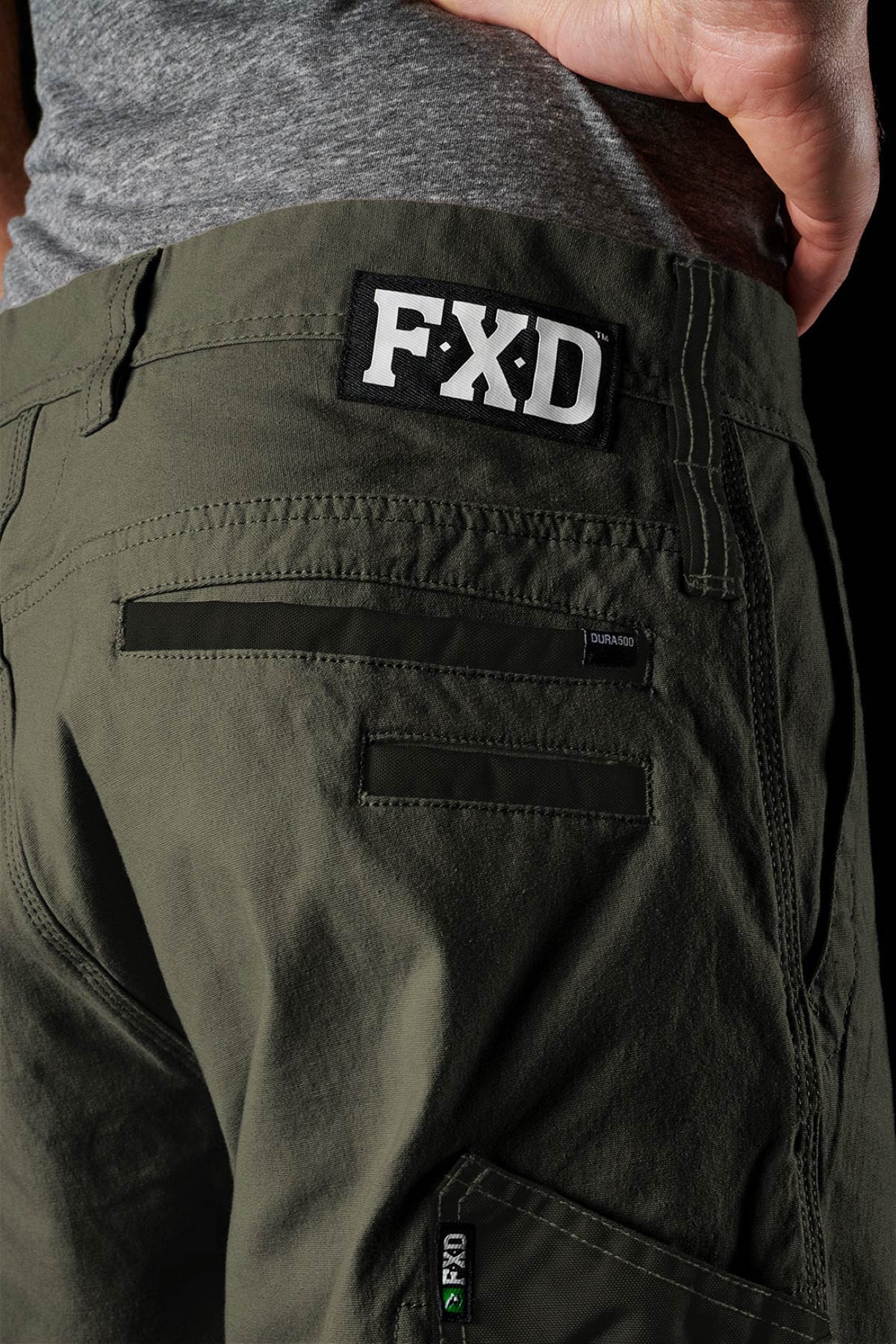 Close-up of a person wearing olive green FXD work shorts and a grey shirt, displaying the FXD logo patch above the back pocket—perfect for fans of comfortable stretch workwear like FXD WP-4 pants.