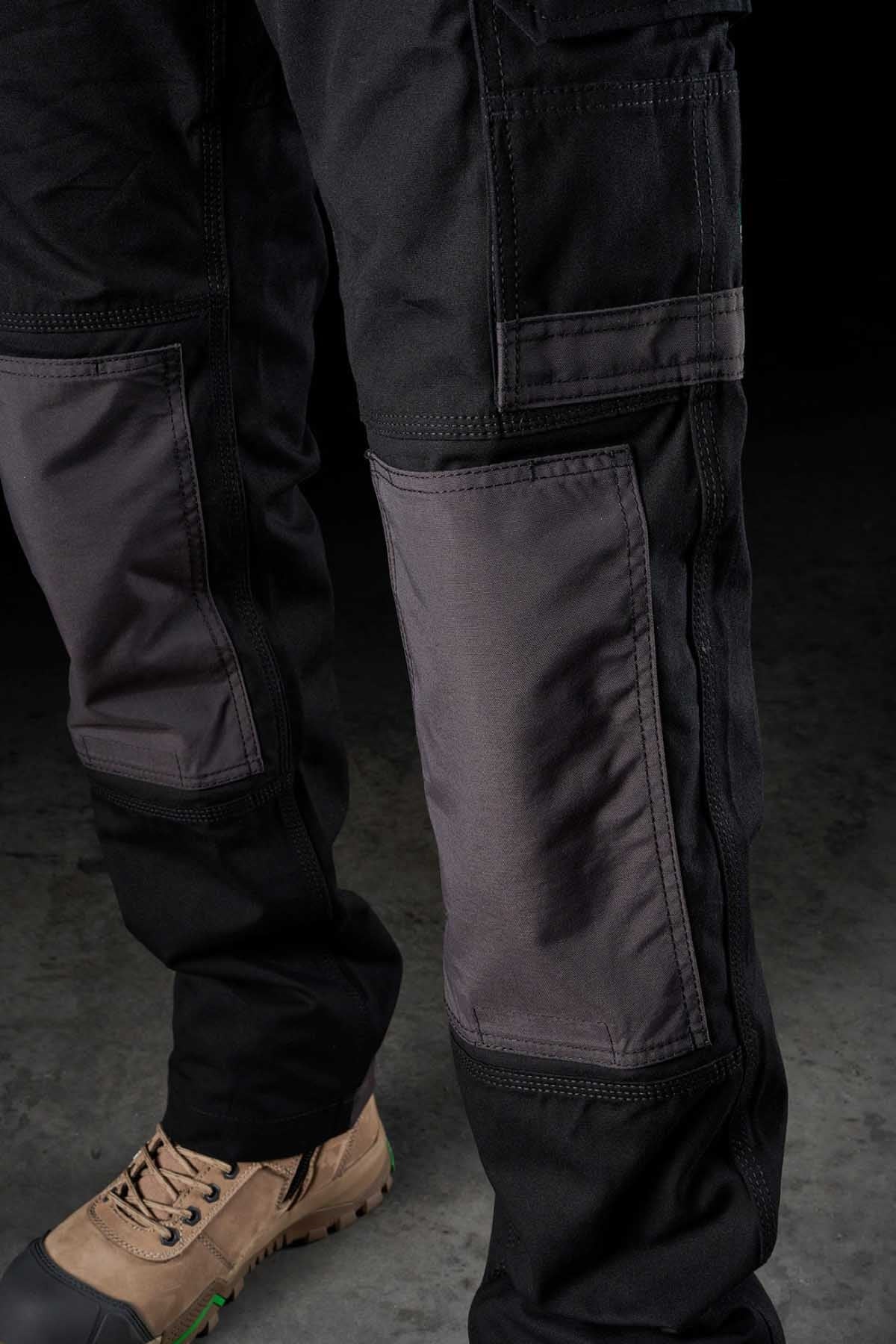A close-up of a person wearing FXD WP-1 black and gray cargo pants featuring reinforced knee patches and Duratech reinforced pockets. The person is also sporting comfortable tan work boots with green-tipped soles. The background is a dark, textured surface.