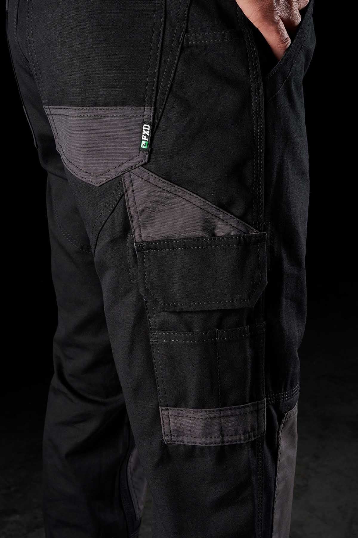Close-up of a person wearing FXD WP-1 pants, black with gray accents. These comfortable work pants feature multiple pockets, including Duratech reinforced pockets and a large side pocket. A label with the text "FIT00" in green and white is visible. The person's hand rests casually in one of the front pockets.