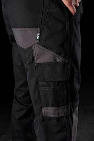 Close-up of a person wearing FXD WP-1 pants, black with gray accents. These comfortable work pants feature multiple pockets, including Duratech reinforced pockets and a large side pocket. A label with the text "FIT00" in green and white is visible. The person's hand rests casually in one of the front pockets.