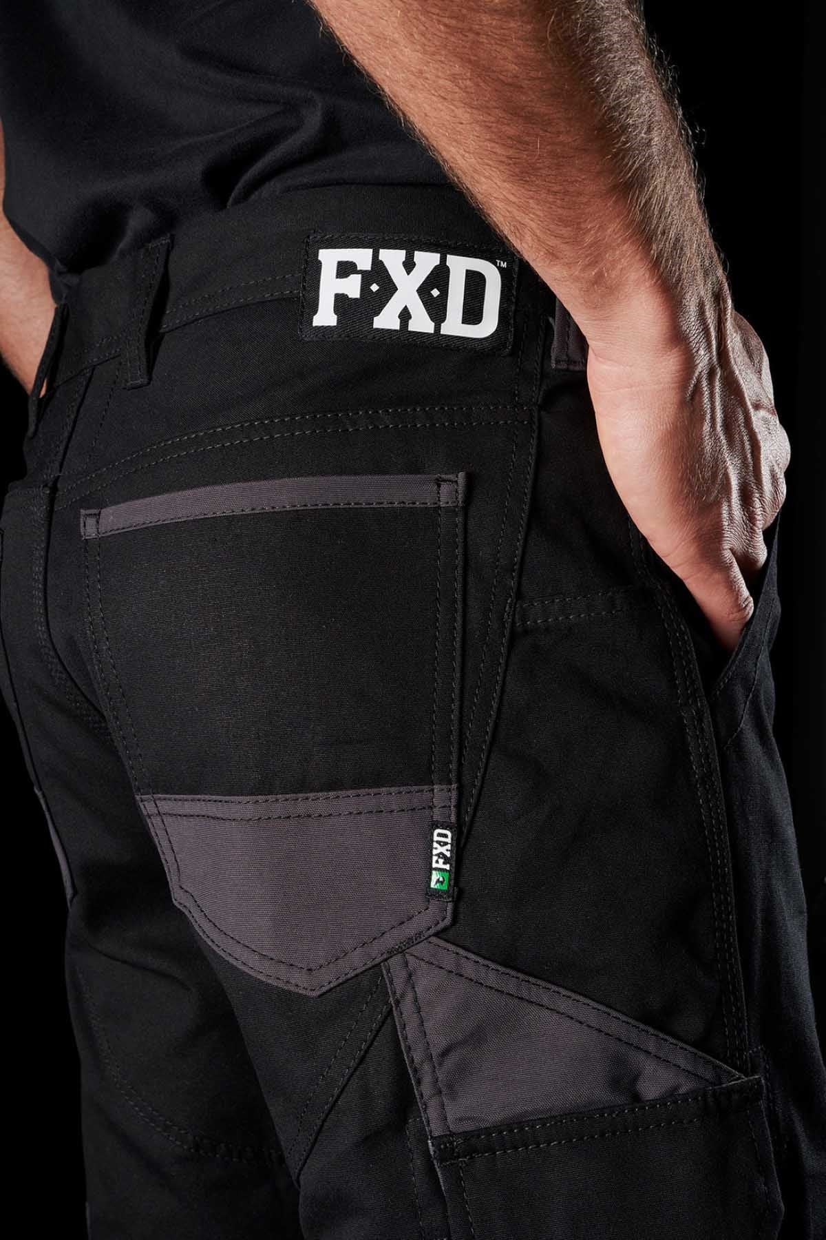 An individual is seen wearing black FXD WP-1 work pants, highlighting the side pocket design with Duratech reinforced stitching. These comfortable work pants boast multiple pockets and an FXD logo patch above the back pocket. The image emphasizes the pants' 360-degree stretch and fine details against a dark background.