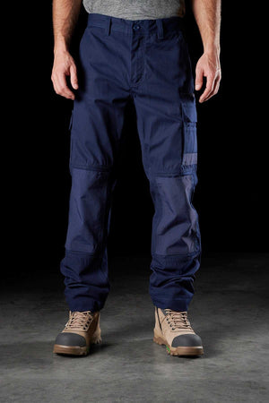 A person stands against a dark background wearing FXD WP-1 comfortable work pants in navy blue, featuring Duratech reinforced pockets and multiple sections. The pants have reinforced knees and are paired with tan work boots. The person's torso is not visible, and their arms are slightly spread out.