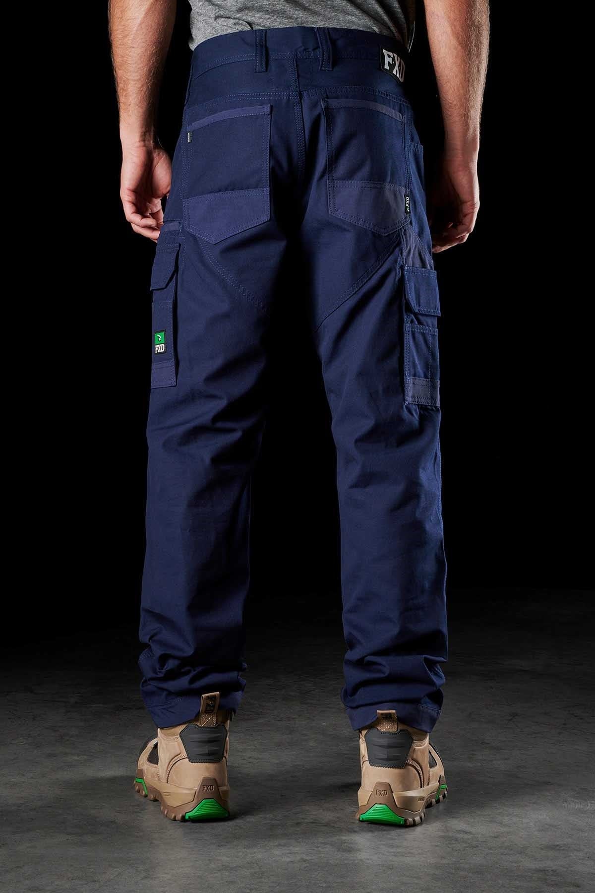 A person stands with their back to the camera, showcasing the FXD WP-1 cargo pants in dark blue, featuring Duratech reinforced pockets and multiple compartments. They are wearing tan work boots with green accents. The dark background highlights the comfortable work pant and footwear.