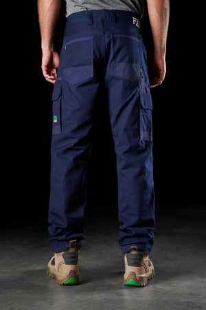 A person stands with their back to the camera, showcasing the FXD WP-1 cargo pants in dark blue, featuring Duratech reinforced pockets and multiple compartments. They are wearing tan work boots with green accents. The dark background highlights the comfortable work pant and footwear.