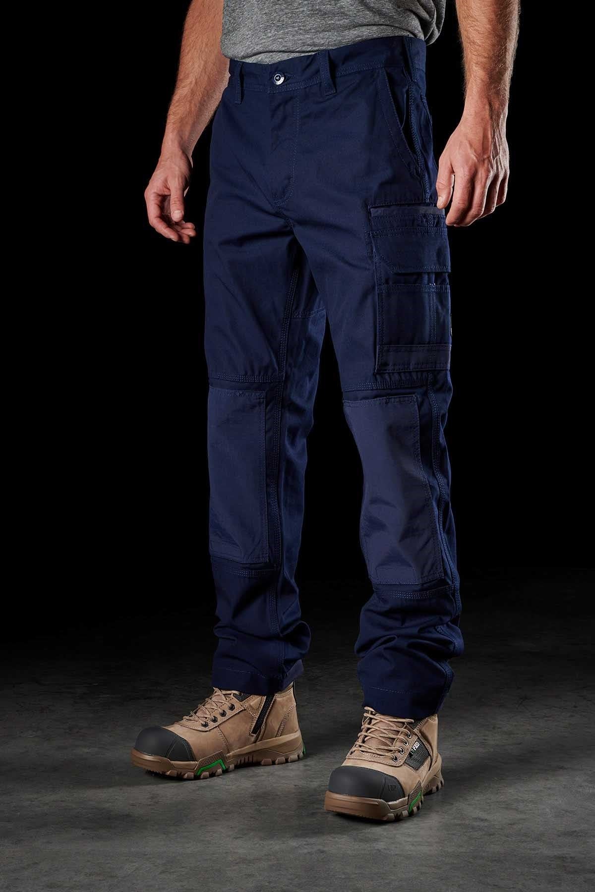 A person wearing FXD WP-1 dark blue cargo pants with multiple Duratech reinforced pockets and a gray t-shirt stands against a black background. They are also wearing tan work boots with green accents on the soles. The focus is on the lower half of the body.