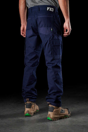 A person is standing with their back to the camera, wearing FXD WP-1 navy blue work pants with Duratech reinforced pockets and a belt reading "FXD." They are also sporting tan hiking boots with green soles. The background is dark and indistinct.