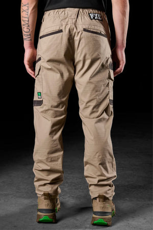 A person stands facing away in FXD WP-12 tan 2-way stretch ripstop pants with utility pockets, black zippers and logos, brown shoes with green soles, and a large left forearm tattoo. The background is dark.