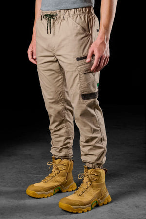 Someone stands on a dark surface wearing FXD WP-12C beige stretch ripstop work pants with black zipper pockets and tan lace-up work boots, arms relaxed by their sides.