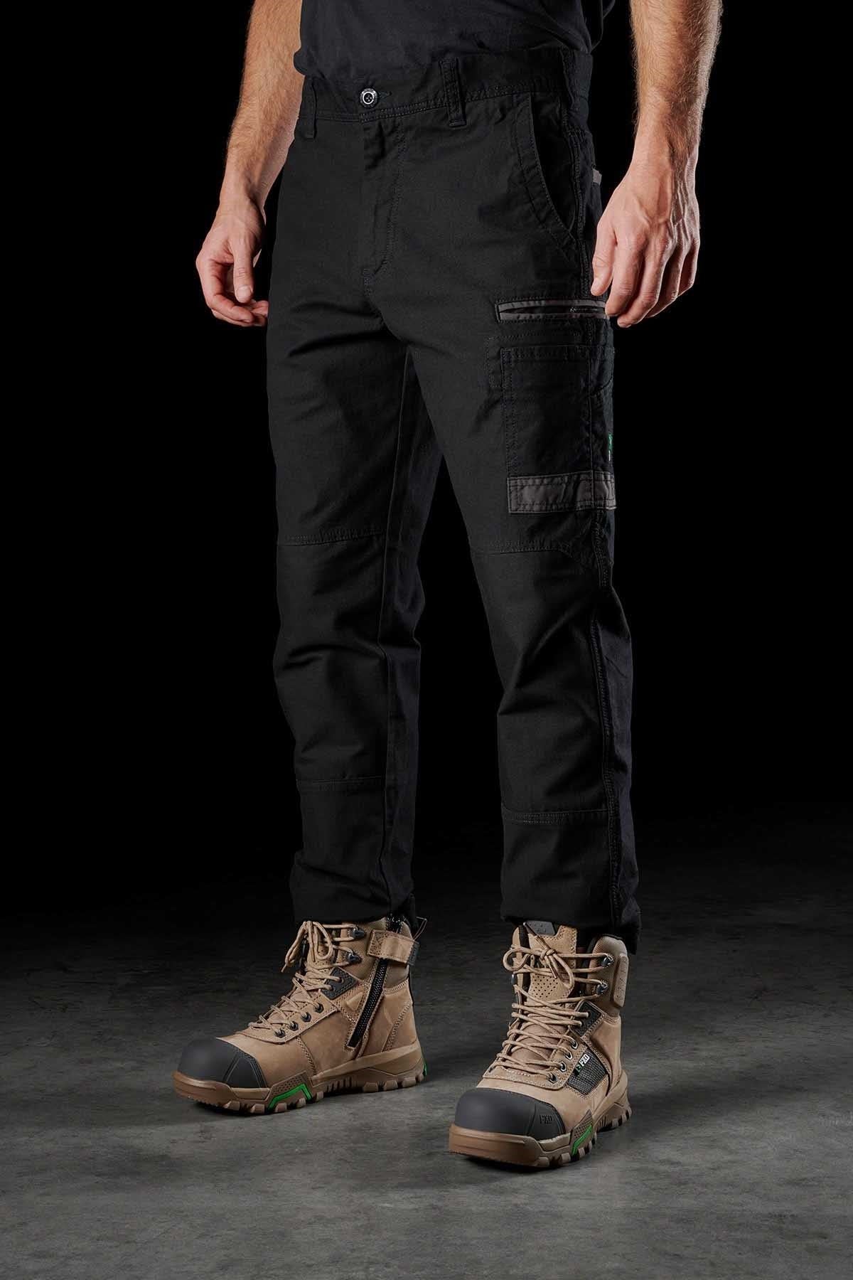 A person wearing FXD WP-3 black stretch cotton cargo pants with reinforced pockets and brown lace-up work boots stands against a dark background. The image focuses on the lower half of their body, showcasing the detailed design of the FXD WP-3 work pant and the rugged, sturdy boots.