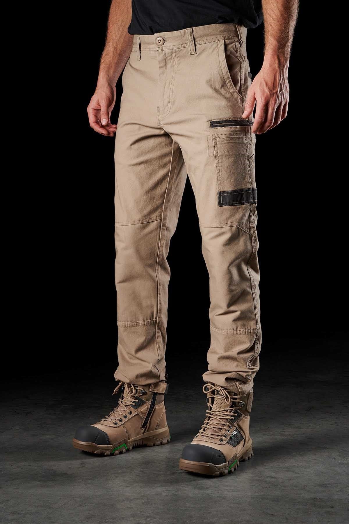 A person wearing FXD WP-3 beige cargo work pants featuring reinforced pockets and black detailing, paired with sturdy brown hiking boots. The person is standing on a gray surface against a black background. The sleeves of a black shirt are partially visible.