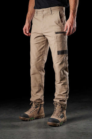 A person wearing FXD WP-3 beige cargo work pants featuring reinforced pockets and black detailing, paired with sturdy brown hiking boots. The person is standing on a gray surface against a black background. The sleeves of a black shirt are partially visible.