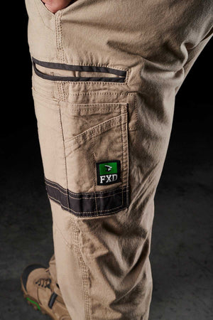 Close-up view of a person wearing FXD WP-3 khaki work pants with black accents and an "FXD" logo on the side pocket. The focus is on the reinforced pockets, showcasing detailed stitching and an additional zipper pocket. The person is also wearing tan work boots with green accents.