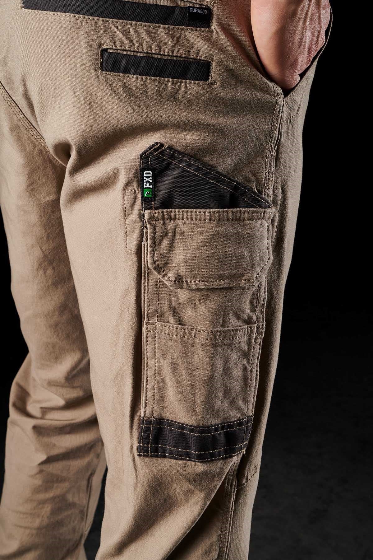 Close-up of a person wearing FXD WP-3 tan work pants, featuring reinforced pockets with black trim and a "Dynamico" label. The hand rests partially in the back pocket, emphasizing the 360-degree stretch material. The background is black.