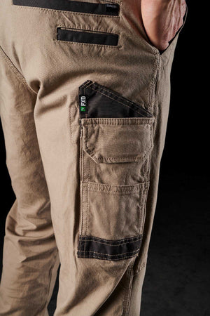 Close-up of a person wearing FXD WP-3 tan work pants, featuring reinforced pockets with black trim and a "Dynamico" label. The hand rests partially in the back pocket, emphasizing the 360-degree stretch material. The background is black.