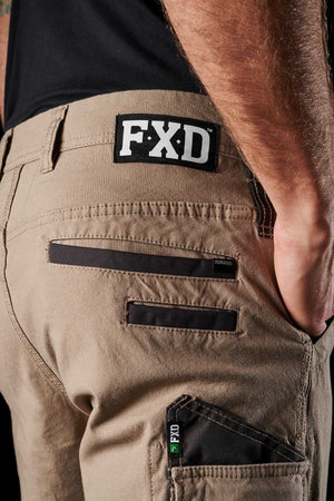 Close-up of a person wearing FXD WP-3 work pants in light brown, featuring reinforced pockets and black patch details. The brand name "FXD" stands out on a black patch near the waistband. Designed with 360-degree stretch, these work pants provide exceptional comfort and flexibility. The person is wearing a black shirt; only the lower back and upper leg are visible.