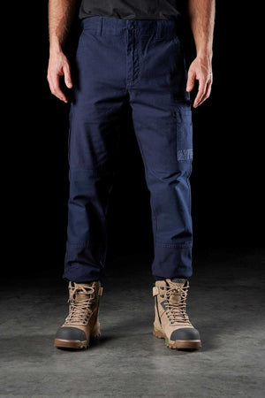 A person is standing in a dim environment, dressed in navy blue cargo pants and tan work boots. The focus is from the waist down, emphasizing the FXD WP-3 Cotton work pant that provides 360-degree stretch. Their hands are resting on the sides of their legs.