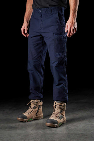 Against a dark background, a person stands wearing FXD WP-3 navy blue cotton cargo pants with multiple pockets and rugged, tan lace-up work boots. The individual is also sporting a black shirt, though only the lower torso and legs are visible in the image.