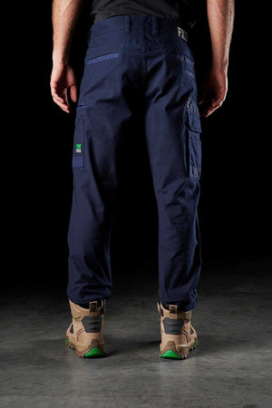 A person wearing FXD WP-3 work pants in dark blue Formatik Cotton stands with their back to the camera. They are sporting tan work boots with green accents. The background is black, drawing attention to the lower half of the person’s body and highlighting the details of both the pants and boots.