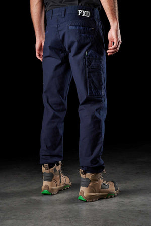 A person stands facing away, showcasing FXD WP-3 navy blue cargo pants made from Formatik Cotton. The work pants feature multiple pockets, the FXD logo on the waistband, and 360-degree stretch for added comfort. The person also wears brown work boots with green accents, set against a dark background.
