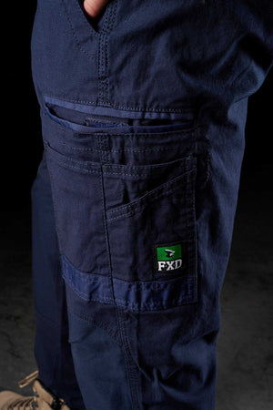 Close-up of a person wearing FXD WP-3 navy blue work pants with multiple pockets and an FXD logo patch on the side pocket, made from durable Formatik Cotton. The background is dark, and only part of the person's legs are visible. They’re also sporting beige work boots.