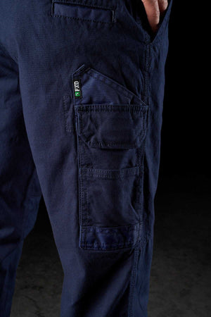 Close-up of a person wearing dark blue FXD WP-3 rugged work pants with multiple pockets. The person has their hand partially inserted into one of the pockets. The background is dark, making the FXD WP-3 Cotton pants with 360-degree stretch stand out prominently.