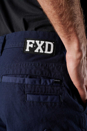 Close-up view of a person wearing FXD WP-3 work pants in dark blue, showcasing an FXD logo patch on the back waistband and a DURASCO label near the rear pocket. Made from Formatik Cotton, these work pants provide 360-degree stretch for optimal comfort and flexibility. The person's arm rests against their hip in the darkened background.