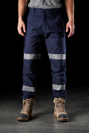 A person stands wearing FXD WP-3T navy blue work pants with 360-degree stretch and reflective tape above and below the knees, paired with tan work boots featuring green accents. The black background highlights the clothing, drawing attention to its functional design. Only the lower half of the person is visible.