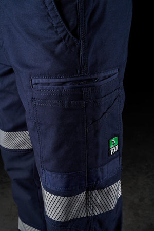 Close-up of a person wearing FXD WP-3T dark blue work pants with reflective silver stripes. The image highlights the side pocket featuring a green and white FXD logo patch. These durable work pants, with their 360-degree stretch and reflective tape, are perfect for environments requiring visibility and safety.