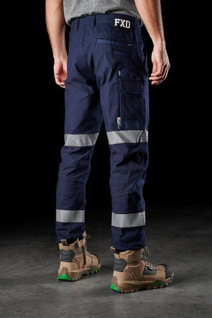 A person wearing dark blue FXD WP-3T work pants with reflective tape and sturdy brown work boots stands facing away. The pants, featuring multiple pockets and the letters "FXD" on the back waistband, offer 360-degree stretch for added comfort. The background is black, highlighting the workwear.