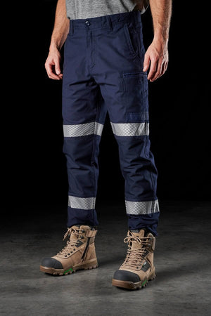 Person wearing FXD WP-3T blue cargo work pants with reflective tape above the knees and sturdy tan boots. The background is dark, highlighting the pants and boots. The person's upper body is mostly out of the frame, with only a portion of a gray shirt visible.