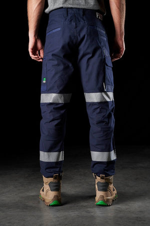 A person is standing against a dark background, wearing FXD WP-3T dark blue work pants featuring reflective tape around the lower legs and side pockets. These pants provide 360-degree stretch for added comfort. The individual is also wearing light brown work boots with green soles. The top portion of their body is out of the frame.