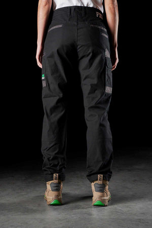 An individual is depicted from the back, donning dark FXD Womens WP-3W work pants with reinforced pockets and patches, along with tan hiking boots featuring green soles. The brand logos and details of the women’s work pants and boots are visible. The setting is a dimly lit area with a concrete floor.