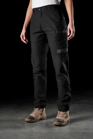 A person wearing FXD Women's WP-3W black cargo pants with multiple reinforced pockets and tan hiking boots stands against a dark background. The work pant, paired with a tucked-in white shirt, offers 360-degree stretch for ultimate comfort. Only the legs and torso are visible, not the head.