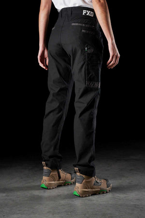 A person wearing a white shirt, FXD Womens WP-3W black utility work pants with reinforced pockets and 360-degree stretch, and brown work boots with green details, stands on a gray floor against a black background. The back of the pants has "FXD" printed on the waistband.