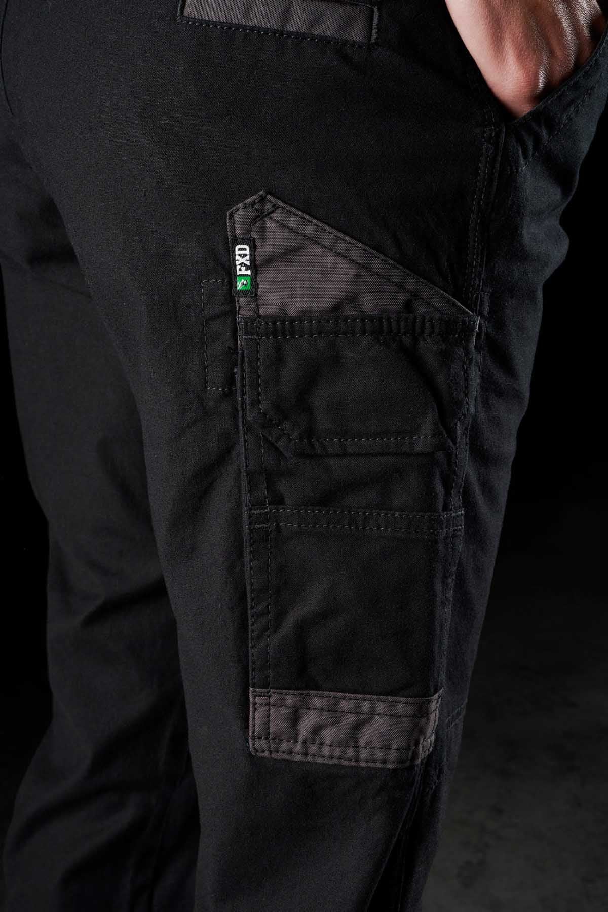 Close-up of a person wearing FXD Womens WP-3W black work pants, featuring a multi-pocket design on the thigh with grey stitching and trims. A small label on the pocket reads "FTD." The reinforced pockets offer 360-degree stretch, while the person's hand is partially visible, resting near the pocket.