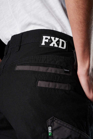 A close-up of a person wearing FXD Womens WP-3W black work pants showcases the visible pockets and stitching details, with the FXD logo prominently displayed on the back waistband. These women's work pants feature reinforced pockets and 360-degree stretch for maximum comfort. The person is also wearing a white shirt tucked into the pants.