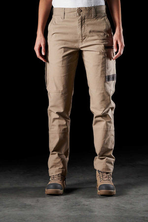 A person is standing against a black background, wearing durable workwear—a beige FXD Womens WP-3W work pant with multiple pockets and brown work boots. The person’s upper body is not visible, only their torso and legs are shown.