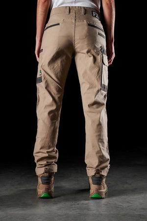 Rear view of a person standing and wearing beige FXD Womens WP-3W work pants with multiple pockets and tan boots. The background is dark, highlighting the durable workwear. The person's torso is partially visible, while details of the pants and boots are in focus.