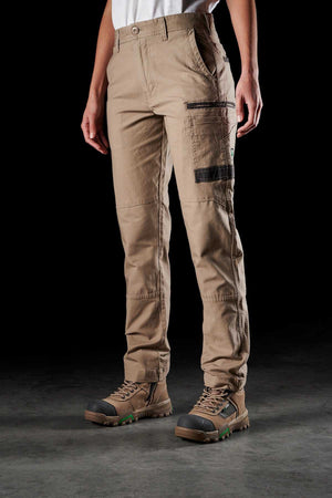 A person is showcasing the FXD Womens WP-3W, a pair of tan cargo pants with multiple pockets and 360-degree stretch, paired with sturdy brown hiking boots. The white shirt contrasts against the black background, emphasizing the durable workwear.